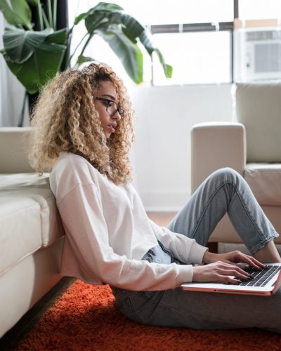 biracial woman with curly blonde hair sitting on floor by white couch, working on a laptop
